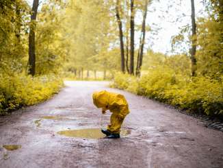 Un enfant porte un imperméable et un pantalon de pluie jaunes ainsi que des bottes en caoutchouc noires. Il joue dans une flaque d'eau sur un chemin forestier.