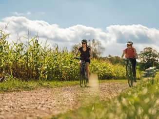 Zwei Freundinnen fahren mit den E-Bikes auf einem Schotterweg durch ein Maisfeld.