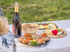 Sur une table recouverte d'une nappe blanche à l'extérieur se trouvent des plateaux avec différentes entrées. Il y a un plateau de fromages, un plateau de pains avec garniture et un plateau de melons.