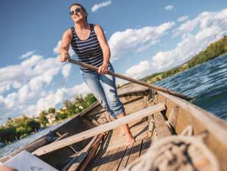 Une femme portant un t-shirt rayé se tient dans un weidling, un long bateau en bois. Dans sa main, elle tient une rame. Elle s'en sert pour diriger le bateau en bois.