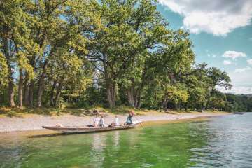 Trois personnes et un chien sont assis dans le weidling, un long bateau en bois. Une femme se tient à l'arrière et pique le long de la rive avec une plage de galets et des arbres.