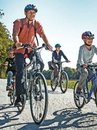 Une famille roule à vélo sur un chemin de terre. Autour d'eux, il y a des prés, des champs et une forêt.