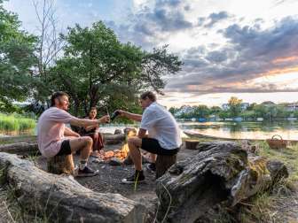 Trois amis sont assis près d'un barbecue au bord du Rhin au crépuscule et trinquent avec une bière.