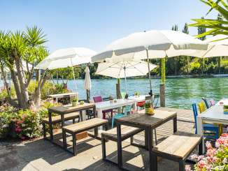 Sur un ponton en bois au bord du Rhin, des tables blanches avec des chaises colorées et des parasols blancs.