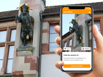 Explorial Schaffhouse Une main tient un téléphone portable devant un bâtiment avec une statue du Bock de Schaffhouse. Sur le téléphone portable, on voit l'image de la statue.