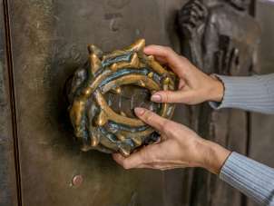 Deux mains palpent la poignée d'une porte d'église. La poignée est un chapelet avec des épines.