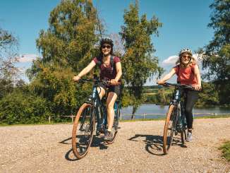 Zwei Freundinnen fahren mit den E-Bikes entlang eines Sees.