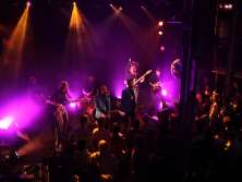 Une pièce sombre, une scène éclairée avec deux chanteurs et cinq autres musiciens. Le reste de la salle est rempli de spectateurs.