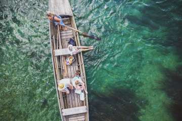 Photo prise d'en haut d'un weidling, un bateau en bois long et étroit. Trois personnes sont assises dans le bateau et une femme se tient à l'arrière et dirige le bateau avec un gouvernail en bois. Le Rhin scintille en vert et bleu.