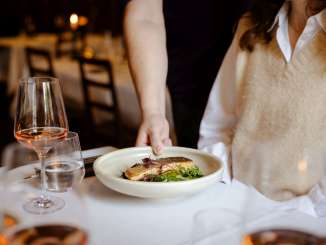 Ein Teller mit einem hübsch angerichteten Fischgericht wird auf einen weiss gedeckten Tisch gestellt. Auf dem Tisch stehen auch mehrere Weingläser mit Rosé.
