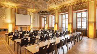 Une salle avec un magnifique plafond en stuc est aménagée pour un séminaire. Devant, il y a un écran et derrière, des rangées de tables avec des chaises.