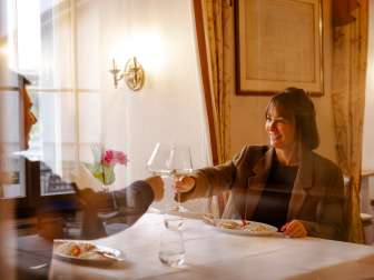 Ein Paar sitz an einem weiss gedeckten Tisch. Sie stossen mit einem Glas Weisswein an und vor Ihnen steht ein Fischgericht.