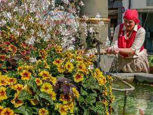 Le roi des fontaines Visite guidée de la fontaine Sur la gauche de la fontaine, des fleurs en fleurs sont visibles. Une dame en jupe brune et haut rouge met les mains dans l'eau de la fontaine. Elle sourit.