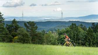 Un homme roule sur un chemin de campagne avec son vélo de gravel jaune. À l'arrière-plan, on aperçoit une vaste étendue de forêts, d'éoliennes et d'un lac.