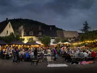 Sur une place, de nombreuses personnes sont assises à des tables en bois. Une guirlande lumineuse crée une ambiance chaleureuse.