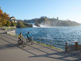 Deux cyclistes au bord d'une rivière. En arrière-plan, on aperçoit une chute d'eau. Sur l'autre rive se trouve un château.
