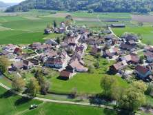 Vue d'en haut sur un petit village entouré de prairies, de champs et de collines boisées.