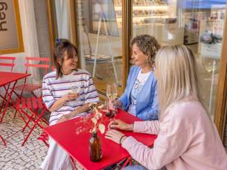 Trois amies sont assises à une petite table de jardin rouge sous une tonnelle dans la vieille ville. Elles ont toutes un verre de vin à la main et bavardent.