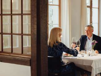 Un couple est assis à une table nappée de blanc. Ils ont un menu gastronomique devant eux et trinquent avec un verre de vin rouge.
