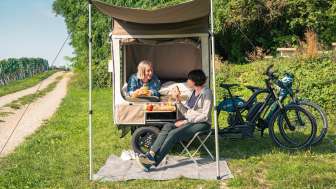 Dans un pré à côté de vignes, il y a un vélo-cargo avec une tente dépliée. Une personne est couchée sur le lit et assise sur une chaise de camping devant.