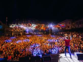 Vue de la scène avec le chanteur sur une place au milieu de la vieille ville remplie de public. Les façades des maisons sont éclairées par un spectacle de lumière.