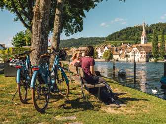 Deux amies sont assises sur un banc en bois au bord du Rhin avec vue sur Stein am Rhein. Derrière elles se trouvent deux vélos électriques.