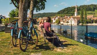 Deux amies sont assises sur un banc en bois au bord du Rhin avec vue sur Stein am Rhein. Derrière elles se trouvent deux vélos électriques.