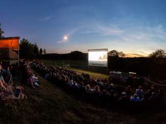 Dans un pré situé sur une petite pente, de nombreuses personnes sont assises et regardent un film sur un grand écran.