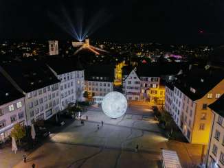 Vue d'en haut, de nuit, sur le Munot illuminé. Des faisceaux de lumière brillent dans le ciel nocturne.