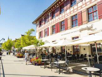 Terrasse Güterhof Schaffhausen La terrasse ensoleillée avec une plateforme en bois, des parasols blancs et des tables avec des chaises noires se trouve directement sur un chemin piétonnier au bord du Rhin.