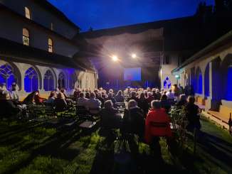 Des sièges sont disposés dans une cour intérieure sur la pelouse. Une scène provisoire est montée. Environ 50 personnes se trouvent dans le public.