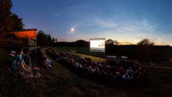 Dans un pré situé sur une petite pente, de nombreuses personnes sont assises et regardent un film sur un grand écran.