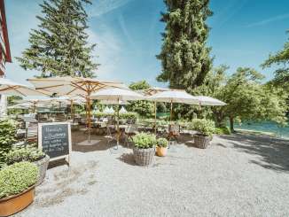 Terrasse de l'auberge Schupfen Des chaises et des tables en bois sont dressées et des parasols blancs offrent de l'ombre. La terrasse du restaurant est située directement au bord du Rhin et entourée de verdure.