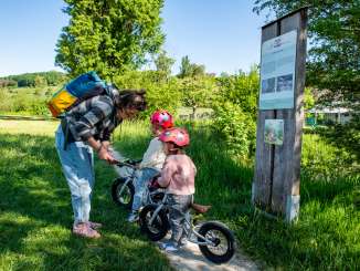 Chemin des castors Thayngen Une femme adulte avec deux enfants sur un sentier près d'une rivière. Les deux enfants sont assis sur une roue d'exercice. A côté se trouve un panneau où l'on peut voir l'image d'un castor.