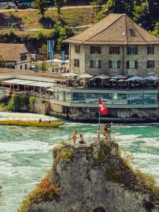Sur le rocher au milieu de la chute d'eau se trouvent plusieurs personnes et un drapeau suisse. Derrière le rocher, on voit le Schlössli Wörth.