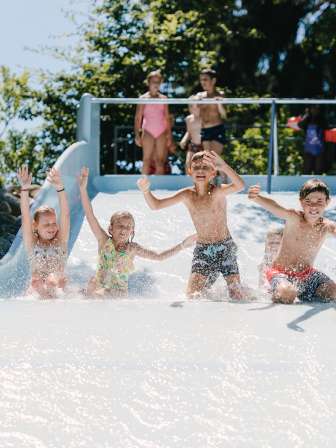 Quatre enfants descendent ensemble un large toboggan dans une piscine extérieure.