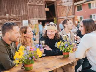 Un groupe d'amis est assis à une table de fête décorée de fleurs. On aperçoit une grange à l'arrière-plan. Les amis trinquent avec un verre de vin.