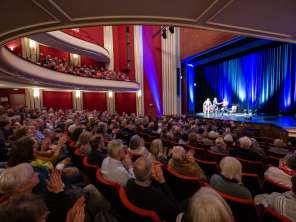 Erzählzeit sans frontières Une salle remplie de gens qui tapent dans leurs mains. Une scène éclairée avec trois personnes dessus, un canapé et un fauteuil.