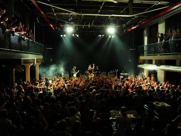 Une salle remplie de gens. Il y a un balcon à gauche et à droite. Devant, il y a une scène avec un groupe qui joue un concert.