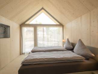 Un lit double avec des draps gris dans une cabane en bois. La grande fenêtre a un rideau qui laisse passer la lumière.