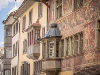 Vue de plusieurs façades de maisons avec un oriel. L'une des façades est légèrement jaune, l'autre richement décorée de peintures.