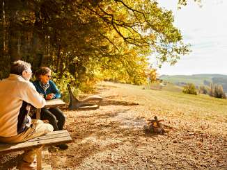 Un couple est assis sur un banc en bois près d'un foyer. La forêt resplendit de ses couleurs automnales.