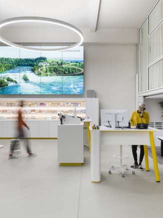 Visitor Centre Schaffhouse C'est la zone des guichets du Visitor Centre de Schaffhouse. Une femme en pull jaune moutarde est debout à une table haute et téléphone. Une autre personne se dirige vers le guichet libre.