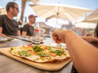 Sur la terrasse d'un restaurant, une tarte flambée aux légumes multicolores est posée sur une table.