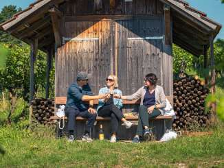 Trois personnes sont assises sur un banc en bois devant un Räbhüüsli dans les vignes. Ils trinquent avec un verre de vin blanc.