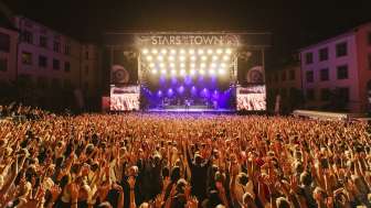 Des stars dans la ville De nombreuses personnes, les bras en l'air, assistent à un concert avec un groupe sur une grande scène.