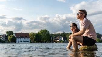 Deux personnes sont assises sur de gros rochers dans le Rhin et se rafraîchissent les pieds dans l'eau.