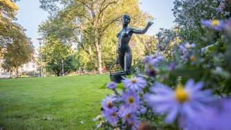 Une statue de femme portant un panier dans un parc. Au premier plan, il y a des fleurs et au second plan, des arbres.