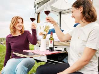 Deux personnes trinquent avec deux verres de vin rouge remplis. Ils sont assis devant une tente de camping.
